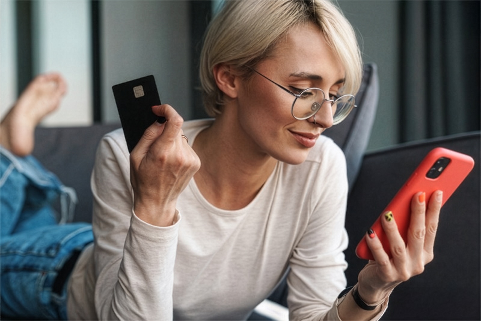 Personne allongée sur un canapé, tenant une carte bancaire dans une main et un téléphone mobile dans l’autre, effectuant un achat en ligne.